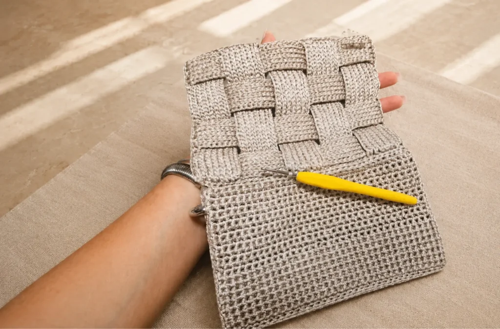 Bolso Dilek de ganchillo hecho a mano en tonos beige sobre mesa de atelier, con antebrazo, dedos y ganchillo amarillo durante el proceso artesanal.
